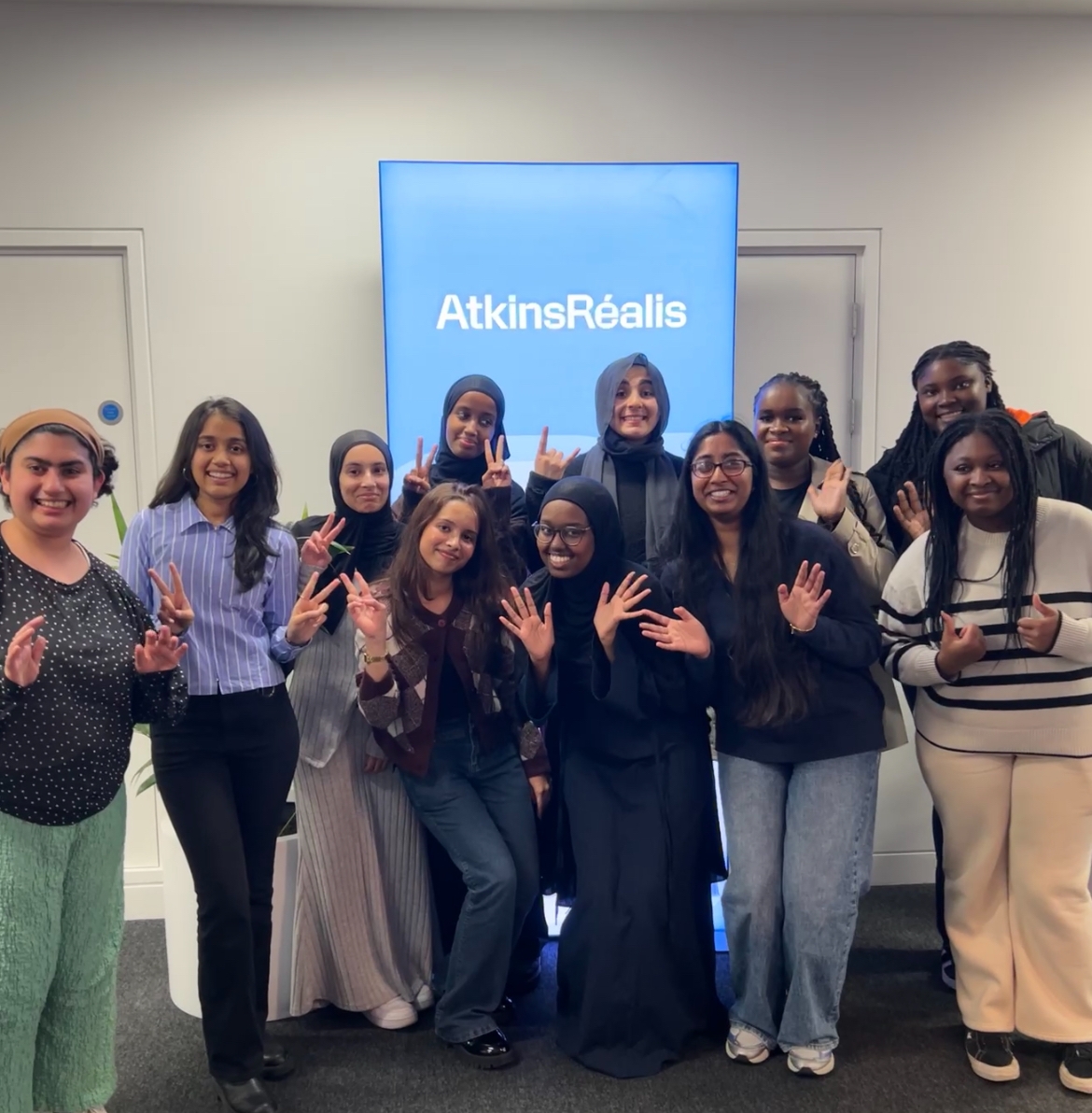 A group of young women stand and crouch in front of an AtkinsRealis sign, smiling and waving.