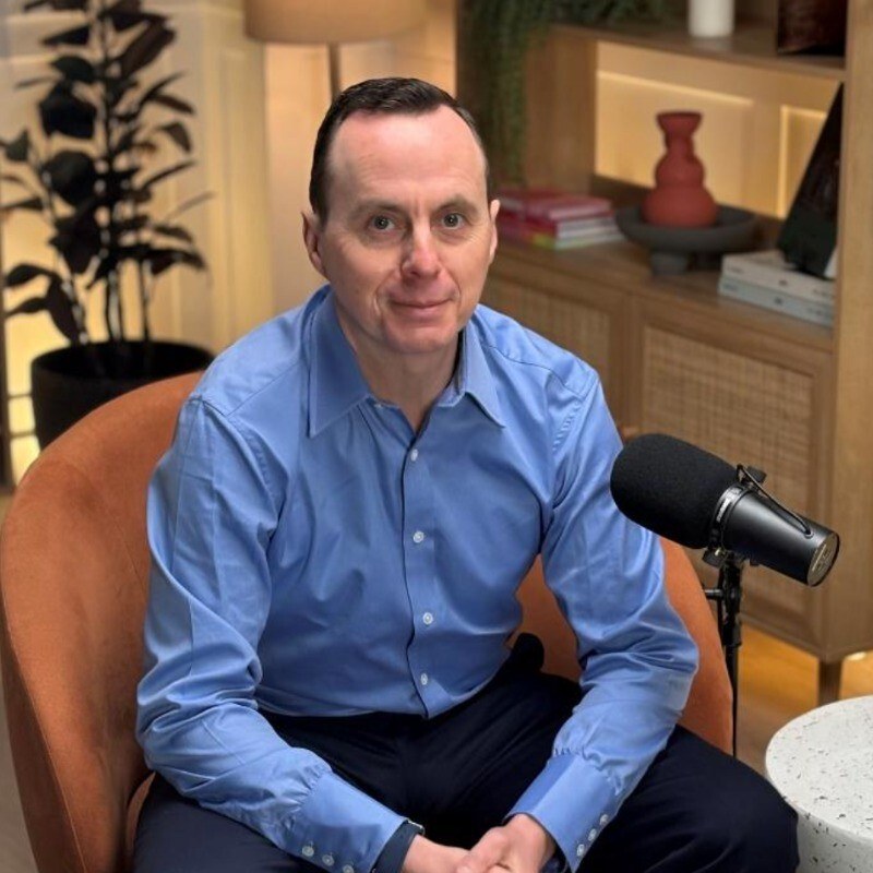 A photo of a white man wearing a blue shirt sat in a chair with a microphone in front of him