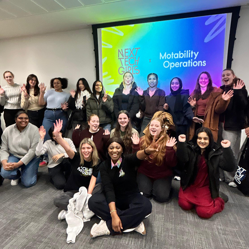 A group of young women smiling and raising their arms in front of a screen that shows the Next Tech Girls and Motability Operations logos, during the Edinburgh hackathon.