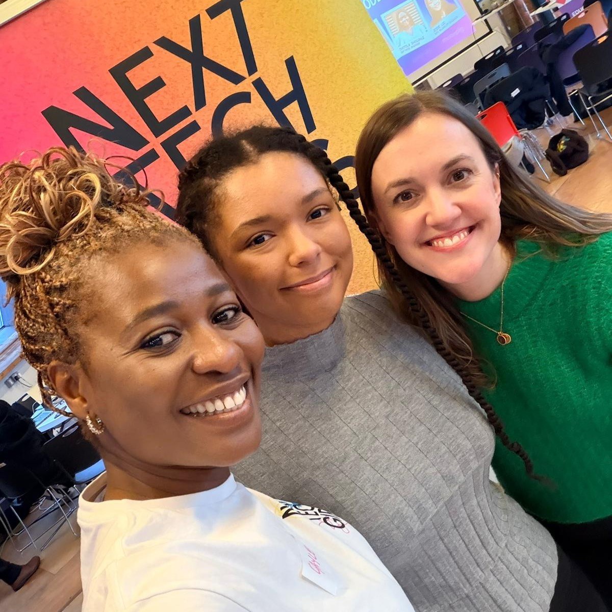 Naomi, an NTG community member, poses and smiles in between NTG team members Emily and Gayle and in front of a colourful Next Tech Girls banner at the Mastek Insight Day in Manchester.