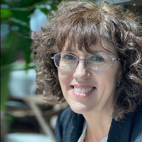 Noel-Ann, a white woman with brown curly hair and glasses, smiles at the camera.