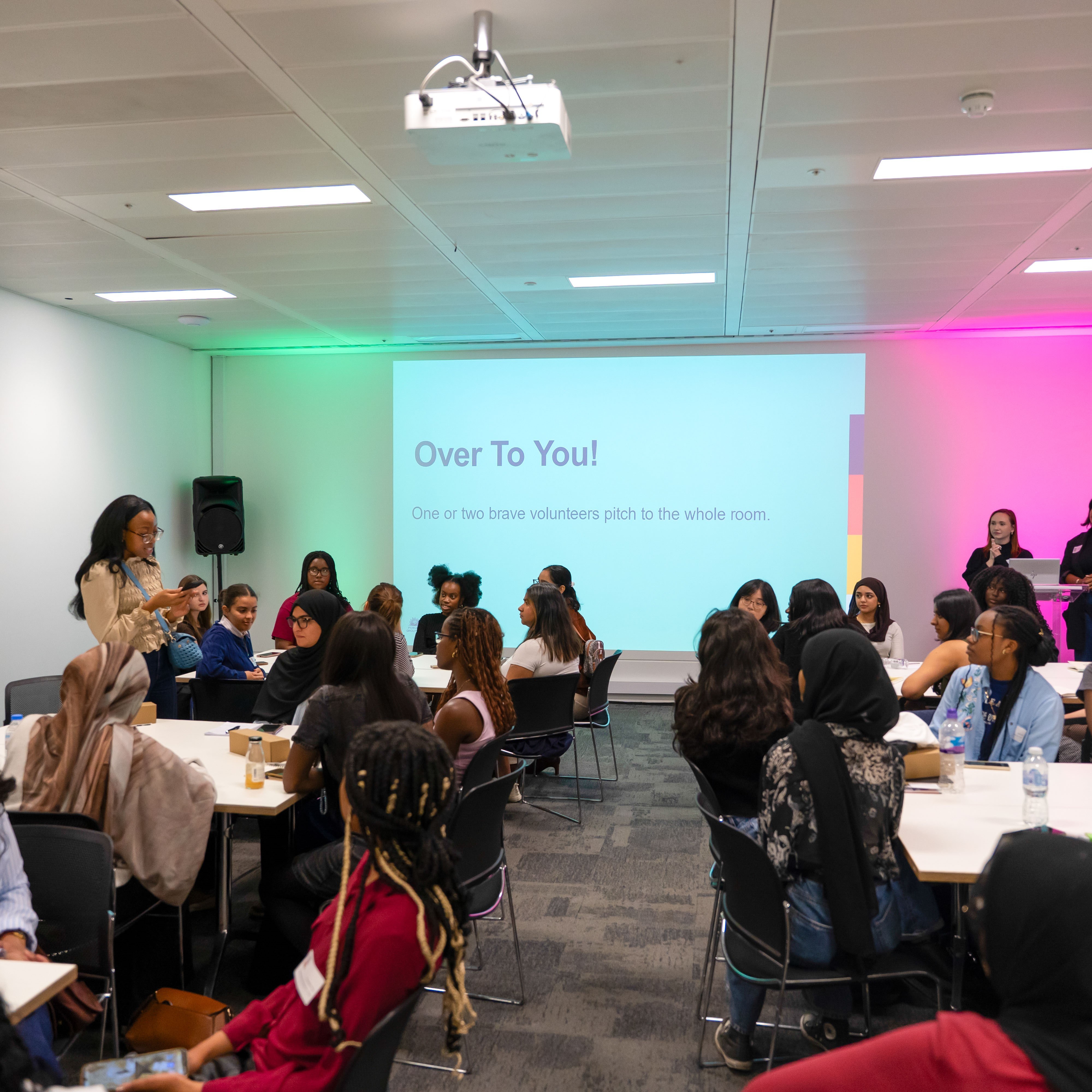 Groups of young women sit round tables in the Publicis Media Pitch Perfect workshop at Next Tech Fest. One woman is standing up reading notes from her phone. There is a screen on the wall at the back of the room that says 'Over to you!'