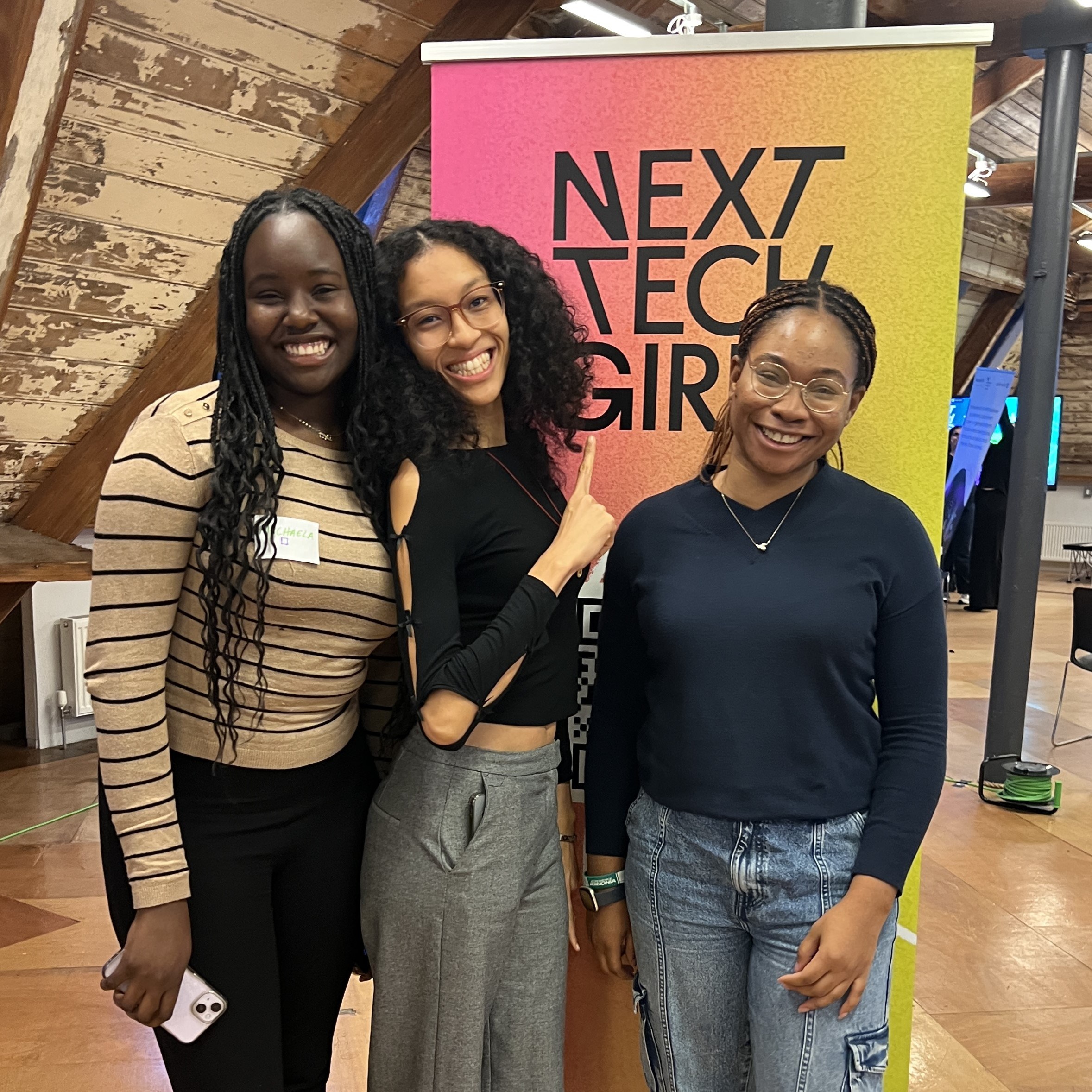 A photo of three young women posing next to a colourful Next Tech Girls banner at the Mastek Insight Day in Manchester.