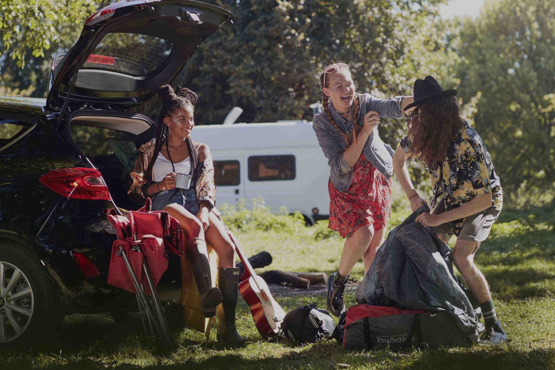 Three festival go-ers in a green area conversing and laughing behind their black car.