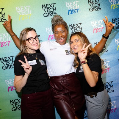 Three women - Emily, Gayle and Mia, the Next Tech Girls team - pose and smile in front of a colourful Next Tech Girls banner