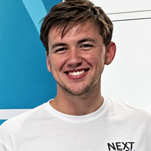 A photo of a young white man in his mid 20s wearing a white Next Tech Girls t shirt and smiling at the camera