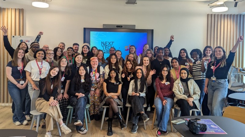 A large group of women who attended the Sky Insight day are standing or sitting for a group picture in a bright indoor space. Many are smiling and waving or holding their arms up.