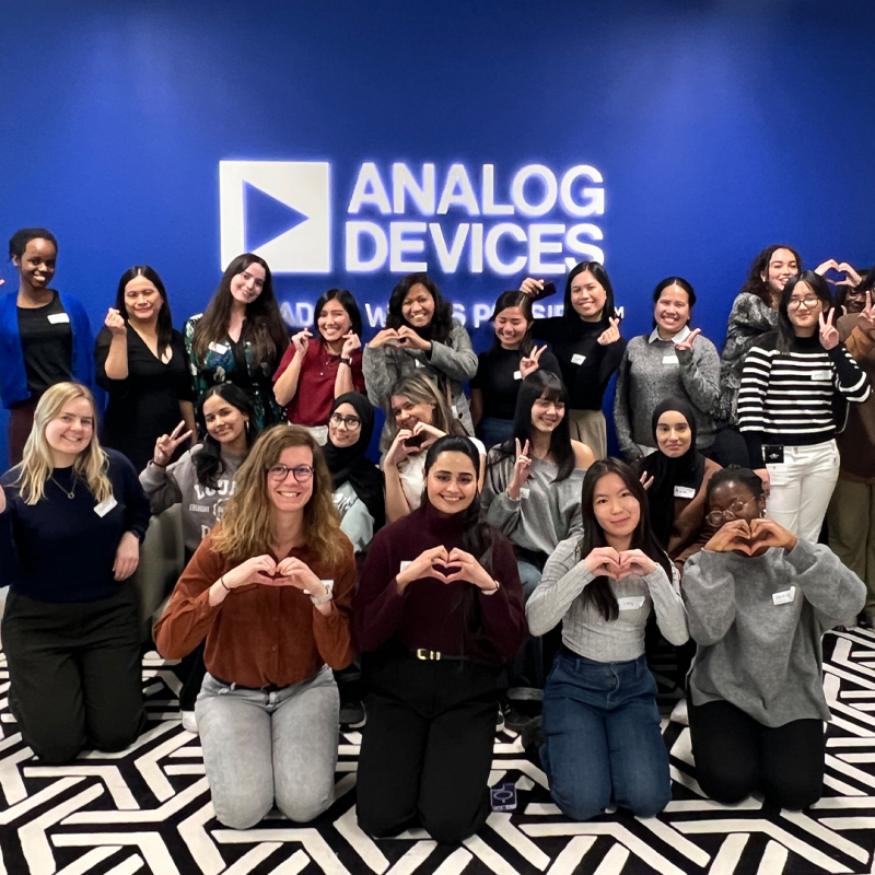 A large group of students and Analog Devices employees - all girls and women taking part in the Next Tech Girls Robotics Insight Day - pose for a group photo, some of them making heart shapes or peace signs with their hands. Behind them is a bright blue wall with the Analog Devices logo on it.