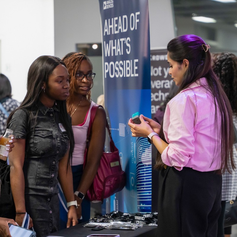 A woman from Analog Devices shows a small technical device to two young women at Next Tech Fest 2025. Behind them is an Analog Devices banner.