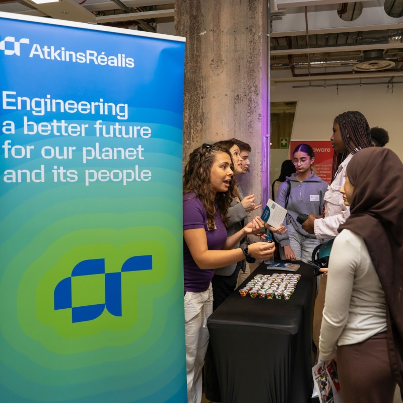 A group of young women crowding round an exhibition stand at Next Tech Fest 2025, speaking to representatives from AtkinsRealis who are stood behind a table and next to a large blue and green AtkinsRealis banner that says 'Engineering a better future for our planet and its people'.