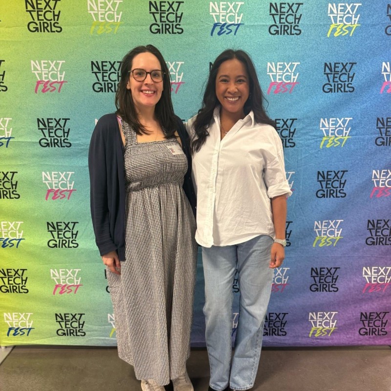 Kate and Venus from Mastek smiling in front of the colourful Next Tech Fest 2025 step and repeat banner.