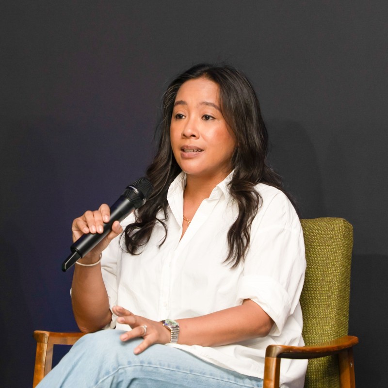 Venus from Mastek speaking on a panel at Next Tech Fest 2025, holding a microphone while seated in a green chair, wearing a white shirt and light blue jeans.