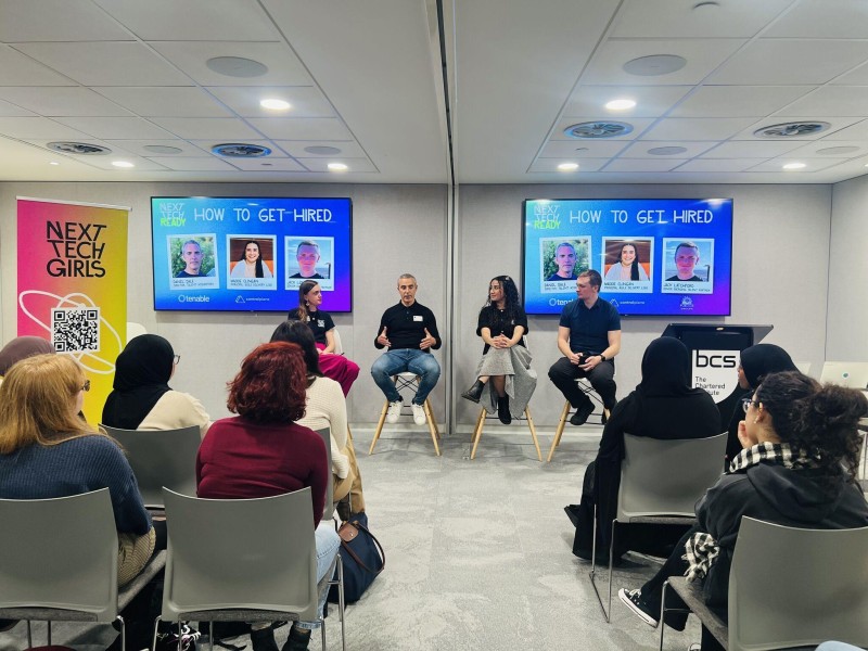 Two men and two women sitting on stools in front of a seated audience of young women during the Next Tech Ready event. Screens either side of them show the panel discussion title is 'How to Get Hired'.