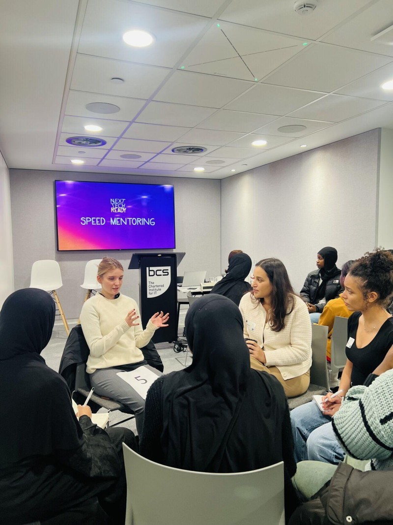 A group of young women sit in a circle, with one talking to the rest of them. A screen behind them says 'Speed Mentoring'.