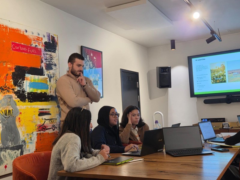 Three young women sit at a table by laptops. A man stands behind them, conducting a workshop as part of their work experience.