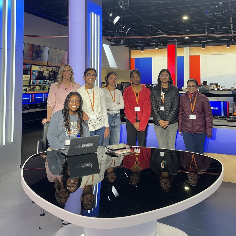A group of young women pose behind a news desk with a Sky Sports News presenter at a Next Tech Girls Insight Day