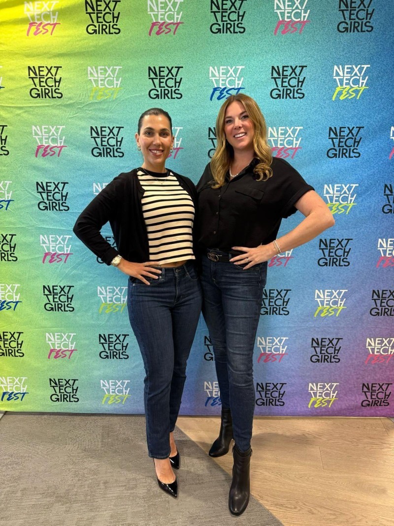 Two women pose in front of a colourful banner that says 'Next Tech Girls' and 'Next Tech Fest' repeated across it.