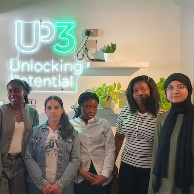 A group of five young women pose and smile in front of a neon sign that says 'UP3 - Unlocking Potential' during their work experience placement
