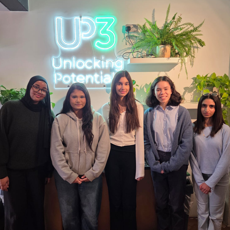 A group of five young women pose and smile in front of a neon sign that says 'UP3 - Unlocking Potential' during their work experience placement