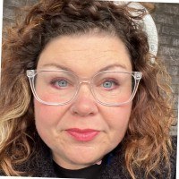 A photo of Vicki Evans. A middle aged white woman with curly reddish hair, blue eyes, and clear framed glasses.